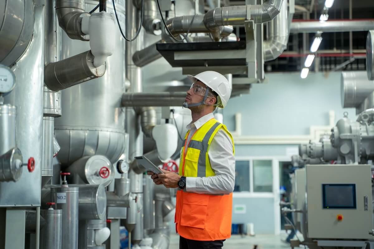 An engineer wearing a white hard hat, safety goggles, and an orange vest stands in an industrial facility, holding a digital tablet while inspecting tall stainless-steel pipes and machinery.
