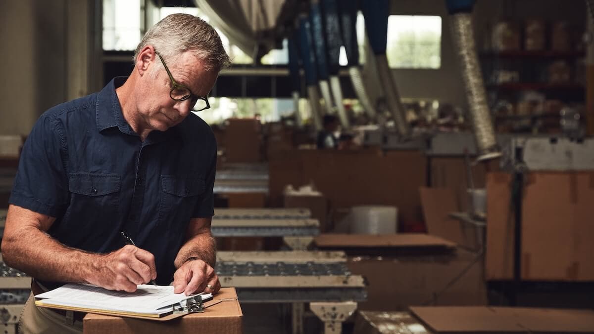 Warehouse supervisor wearing glasses and a navy shirt, standing among packing tables and boxes while writing on a clipboard.